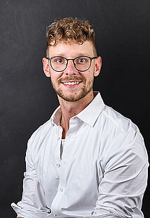 Porträt des lächelnden, jungen Stuckateur-Unternehmers Christian Kast mit lockigen braunen Haaren, Brille und weißem Hemd vor dunklem Hintergrund.