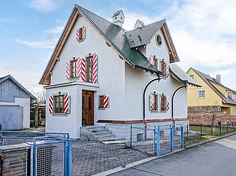 Saniertes, traditionelles Einfamilienhaus mit neuem weißem Außenputz, grünem Dach und dekorativen rot-weiß gestreiften Holz-Fensterläden.