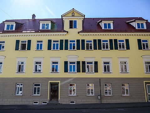 Frontansicht eines sanierten, historischen Mehrfamilienhauses mit gelber Putzfassade, weißen Fensterfaschen, Stuckelementen und dunkelgrünen Fensterläden.