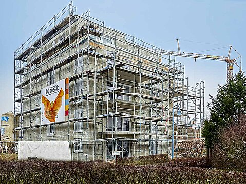 Mehrstöckiges Rohbau-Gebäude komplett eingerüstet, mit großem Werbebanner der Kast GmbH & Co. KG. Kran und Baustelle im Hintergrund.