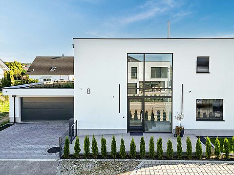 Moderner Neubau mit perfekt ausgeführtem, glattem weißem Außenputz und großflächiger Verglasung im Eingangsbereich. Links eine angebaute Garage mit dunklem Tor.