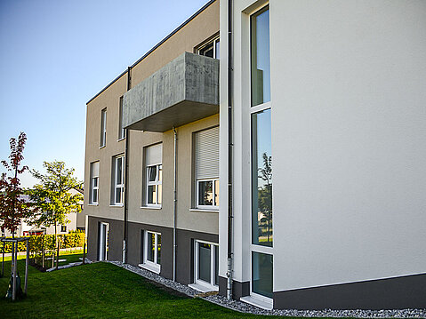 Modern verputztes Wohnhaus mit beiger Fassade, abgesetztem grauen Sockel und Sichtbeton-Balkon.