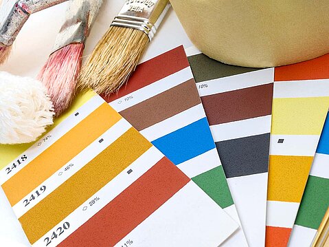 Nahaufnahme von verschiedenen Farbfächern und Farbkarten in leuchtenden Farben, daneben Pinsel und ein Farbtopf, symbolisiert eine professionelle Farbberatung.