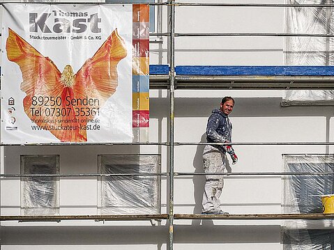 Lächelnder Maler in Arbeitskleidung auf einem Gerüst vor einer weißen Fassade, neben einem Werbebanner der Kast GmbH & Co. KG.