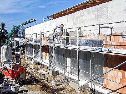 Stuckateur auf einem Arbeitsgerüst beim maschinellen Verputzen einer Ziegelfassade, mit einer roten Putzmaschine im Vordergrund.