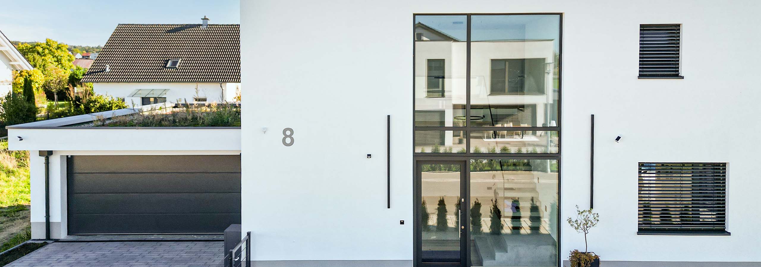 Ausschnitt eines modernen, minimalistischen Hauses mit weißem Außenputz und großflächiger, vertikaler Verglasung im Eingangsbereich, vor blauem Himmel.