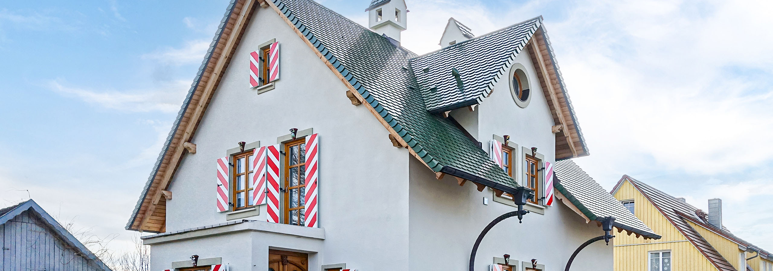 Panorama-Ansicht eines frisch verputzten, weißen Einfamilienhauses im Landhausstil mit grünen Dachziegeln und auffälligen rot-weißen Fensterläden.
